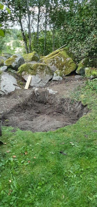 Nygrävd grop i trädgården med stenar och grönska i bakgrunden, redo för makadam och isolering.