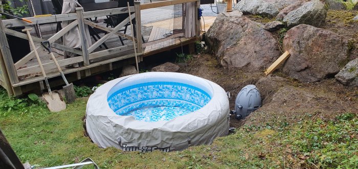 Uppblåsbar pool med vatten intill träveranda och stora stenar, grävmaskiner synliga i trädgården.