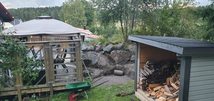 Utsikt över en trädgård med en grushög, en grävd grop och vedskjul med staplad ved, intill en grönskande omgivning.