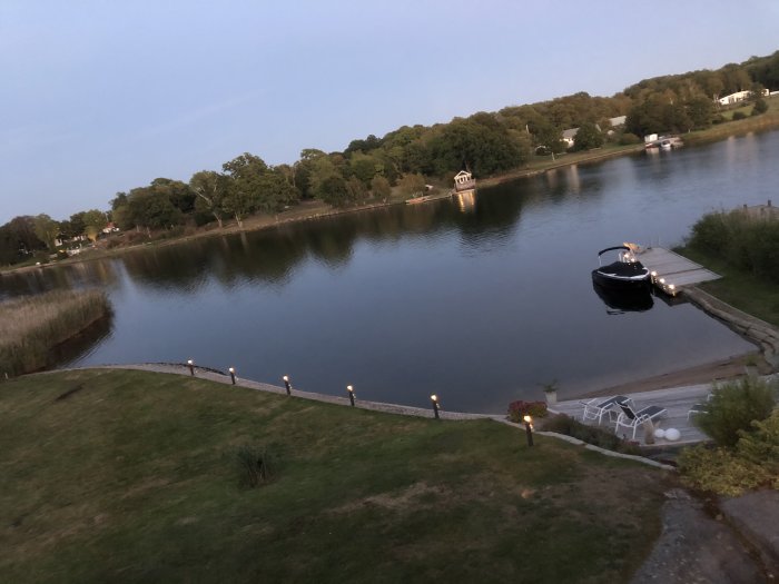 Kvällsutsikt över en stilla sjö med en båt vid bryggan och ljus längs strandkanten.
