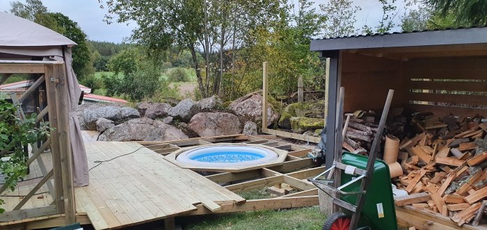 Byggprocess av trädäck runt en rund pool, med omgivande stora stenar och vedskjul fullt med brännved.