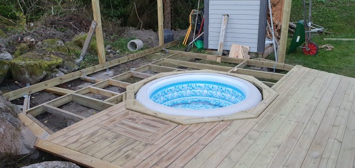 Pågående byggprojekt med trätrall runt en uppblåsbar pool, otäckt plank och soffstomme på en trädgård.