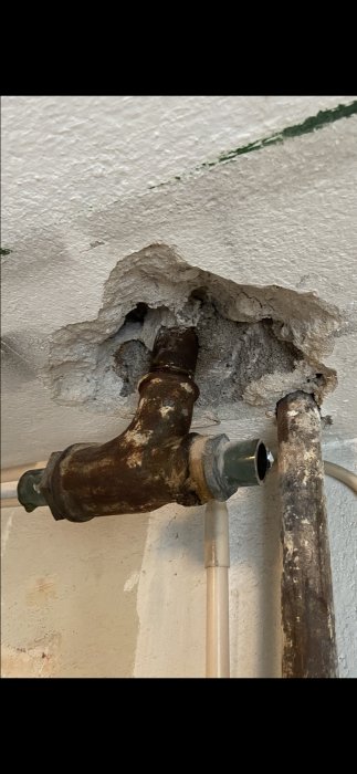 Rostigt rör och koppling synligt genom hål i vit putsad vägg, eventuell värmebehandling planerad.
