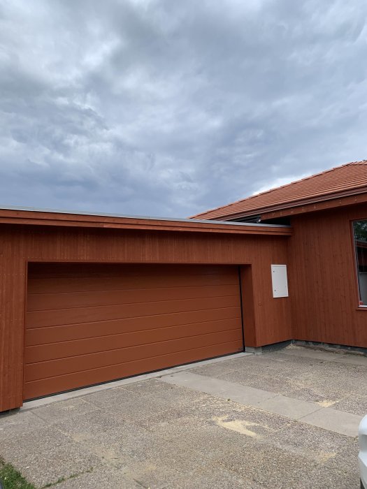 Brun garageport på ett hus med röd träfasad och molnig himmel i bakgrunden.