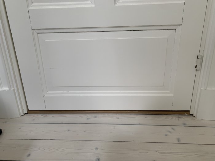 Vit trädörr med en synlig glipa under, ovanför en tröskel på ett trägolv, förberedd för ventilation.