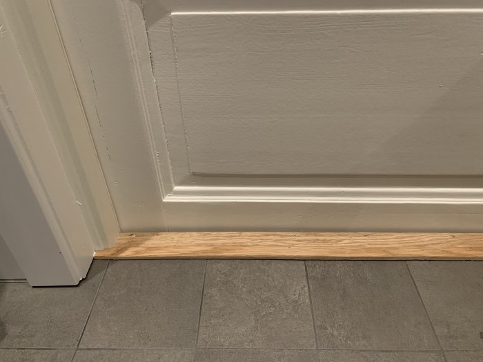 Vit dörr och dörrtröskel i badrum med ofogad karm synlig bredvid grått golv.
