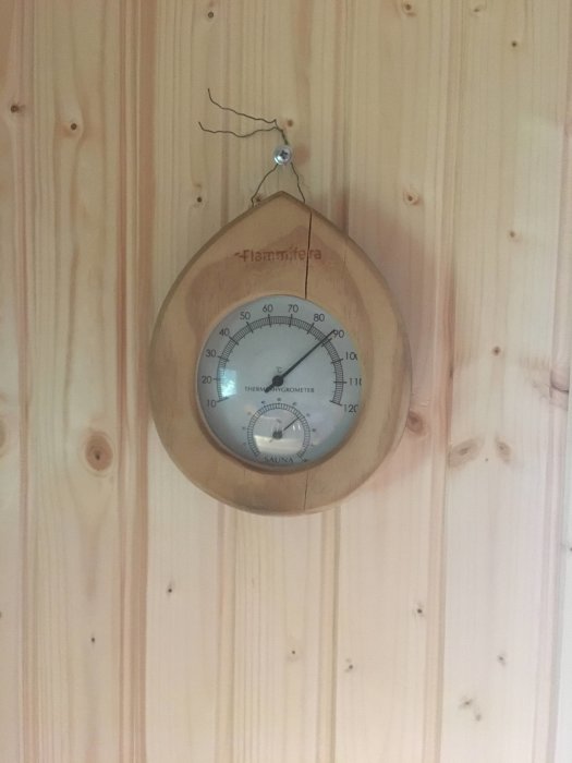 Termometer och hygrometer för bastu upphängd på ljus trävägg, visar cirka 90 grader Celsius.