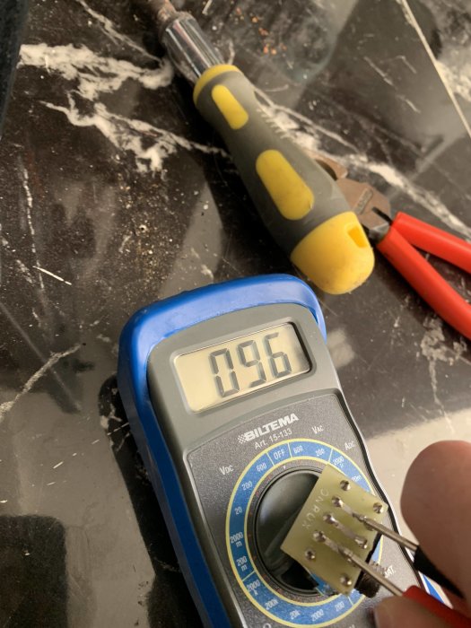 Multimeter visar 0,96 volt med en hand som håller en lödplint, verktyg på en smutsig yta i bakgrunden.