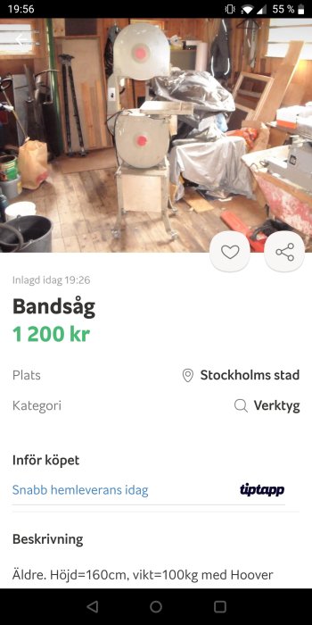 Äldre bandsåg till salu i en rörig verkstad med diverse verktyg och byggmaterial i bakgrunden.