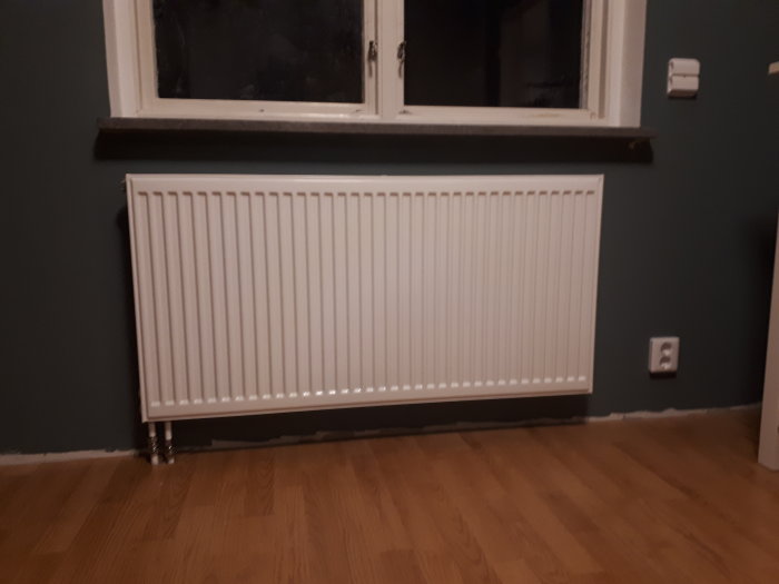 Vit radiator under fönstret med kopplade stål- och kopparrör, färdiginstallerad i ett rum med mörka väggar.