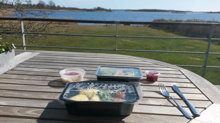 Matlådor på ett utomhusbord med utsikt över en sjö och grönområden i solljus.