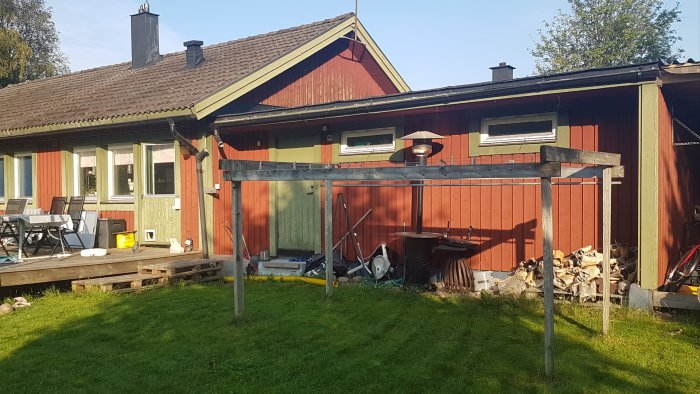 Uppbyggnad av en träkonstruktion utanför ett rött hus, med reglar upphängda på en befintlig byggnad, bredvid ett vedstapel och en trädäck.