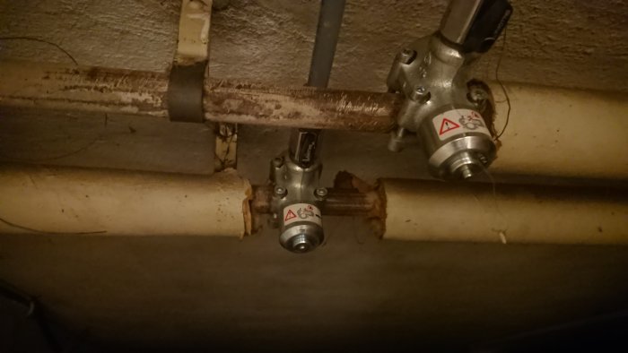 Två termostater monterade på rostiga rörledningar under tak, planerad demontering.