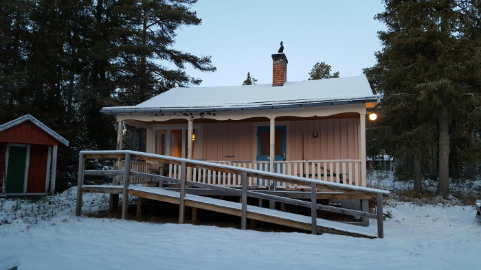 En mysig stuga med snötäckt tak och belysning under en tidig vinterkväll, omgiven av snö och skog.