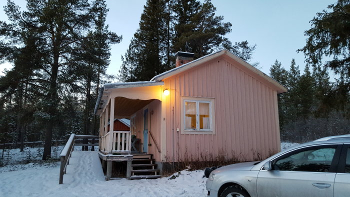 Rosa trästuga med snötäckt tak och mark, öppen veranda och bil parkerad framför, i kvällsbelysning.