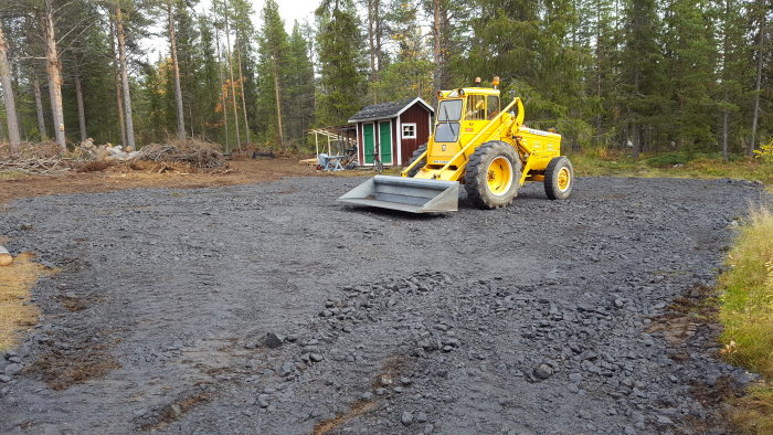 Gult hjullastare jämnar bärlager av krossat material på en blöt byggplats, med skog i bakgrunden.