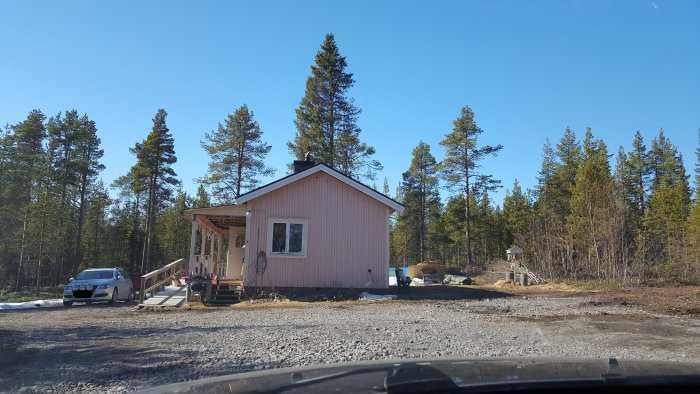 Rosa målat hus med ny panel och gruslagd mark framför, under en klarblå himmel omgivet av tallskog.