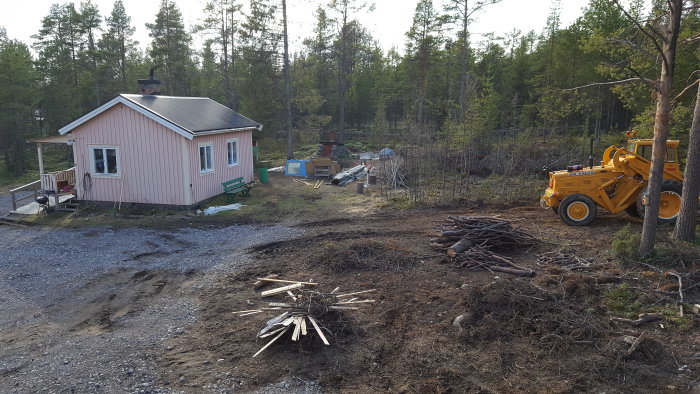 Rosa stuga med nytt tak och veranda, markarbeten framför, vedhögar och en lastmaskin i en skogsmiljö.