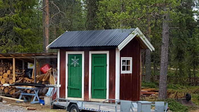 Renoverat skjul med nytt tak och grön dörr, bredvid vedhög och släpvagn i en skogsglänta.