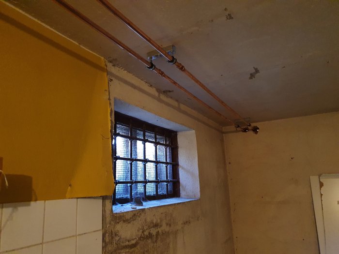 Värmerör installerade längs taket i ett rum under renovering med synligt fönstergaller och gult väggavsnitt.