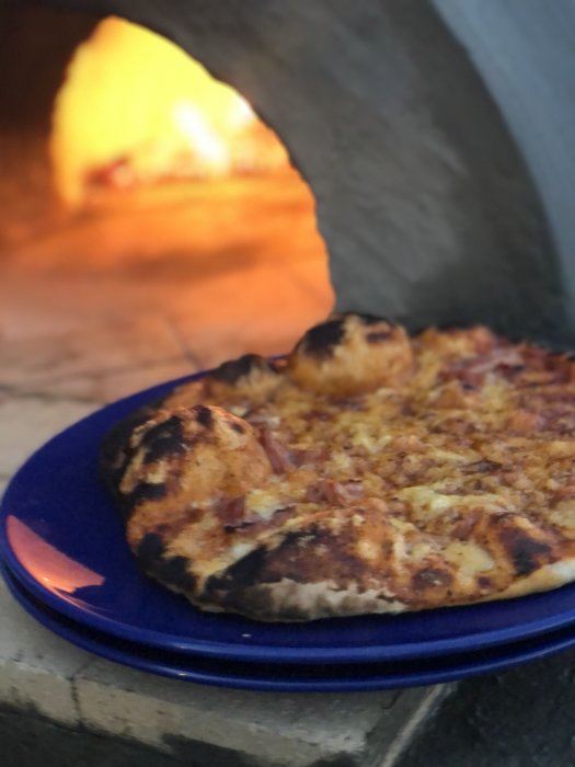 Nygräddad pizza på blå tallrik framför hemmabyggd pizzaugn med eldslåga.