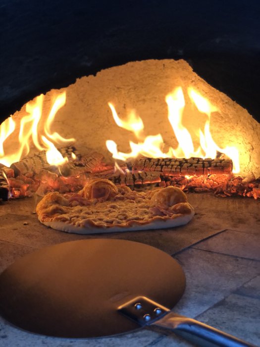 Hembyggd pizzaugn med flammande eld och nygräddad pizza inuti på stenplattan.