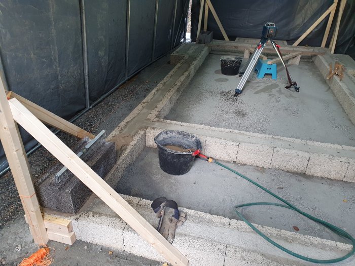 Grundläggning av en källare med betongblock och sand under ett skyddande tält, verktyg och byggmaterial synliga.