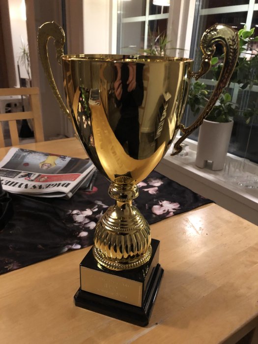 Stor gyllene pokal på ett bord med en reflektion av en person i pokalens glänsande yta.