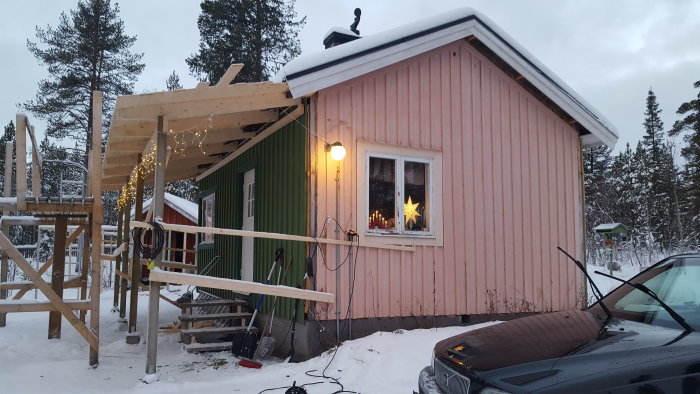 Byggplats med halvfärdigt altantak på vinter, upplyst fönster och juldekorationer, snöigt landskap.