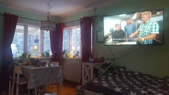 Vardagsrum med matbord belamrat med saker och TV på väggen, utsikt till snöigt landskap genom fönstret.