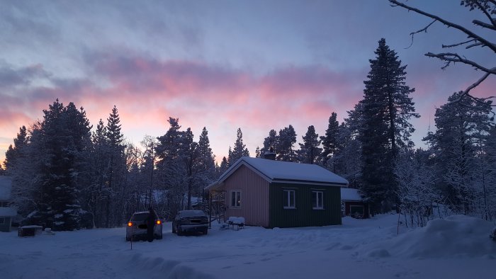 Stuga i vinterskrud med snötäckt landskap och pastellfärgad himmel vid gryning.