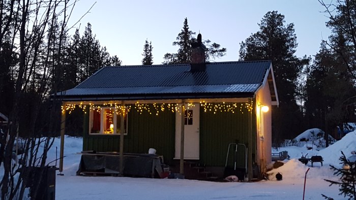 En grön stuga med julbelysning och täckt av snö under en vinterkväll.