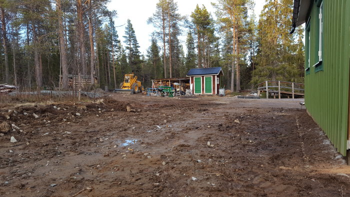 En schaktad baksida med lös jord framför, en grön uthusbyggnad till höger och en gul grävmaskin till vänster i skogsmiljö.