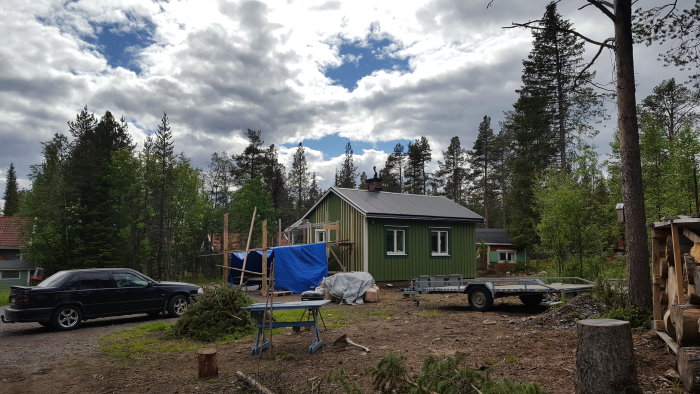 Renoveringsprojekt: Grönt hus med tillbyggnad och byggtält, bilar och släpvagn på förgrund i skogstomt.