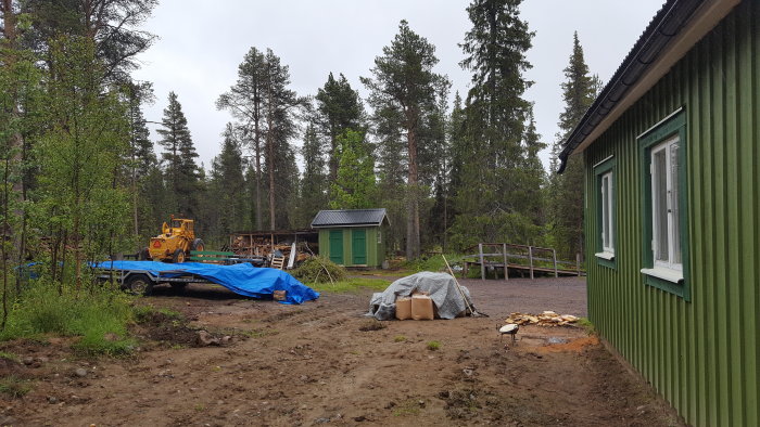 Schaktning på gård med baklastare, grönt hus och uthus, byggmaterial under presenning i skogsmiljö.