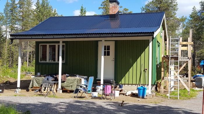 Renoveringsprojekt av en grön stuga med svart tak, skorsten och en stegar vid gaveln, verktyg och en ekorre i förgrunden.