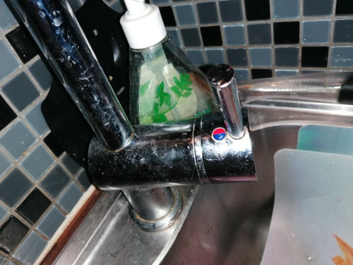 Kökskran med kalkavlagringar och mörk kakelbakgrund, bredvid en diskmedelsflaska.