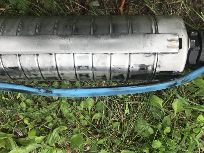 Skadad vattenpump med avskalad blå kabel och repor på metallhöljet, liggande i gräset.