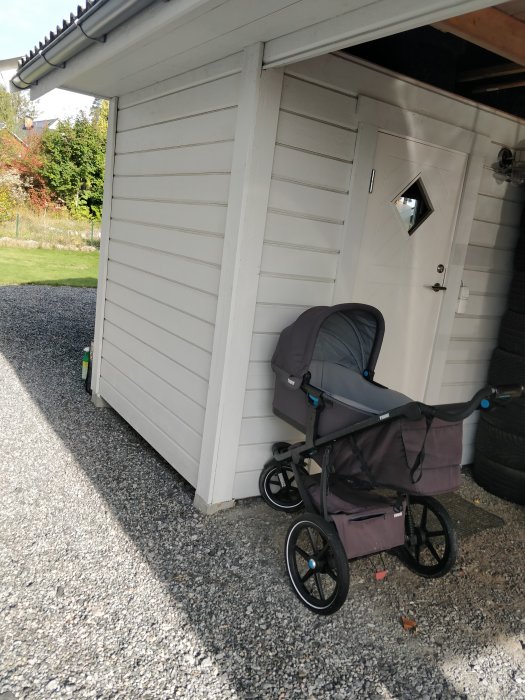 Carport med vit träbeklädnad och förrådsdörr, barnvagn framför och plintar synliga.