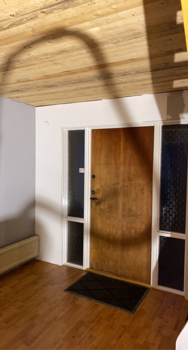 Hall med trägolv och dörr, påbörjad tapetsering på vägg och träbjälkar i taket.