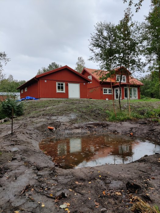 Nygrävd damm med vattenspegel framför ett rött hus med gröna träd i bakgrunden.
