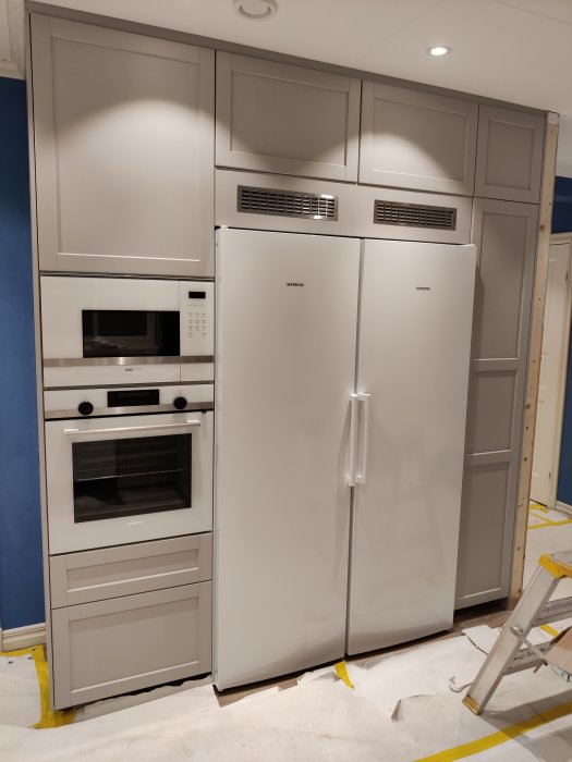Nyinstallerade vitvaror i ett kök under renovering med mikrovågsugn, ugn, kylskåp och inbyggda skåp.