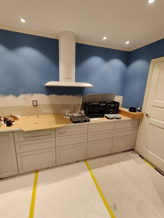 Kök under renovering med oavslutad blå väggmålning, vit köksinredning och fläkt utan kakel.
