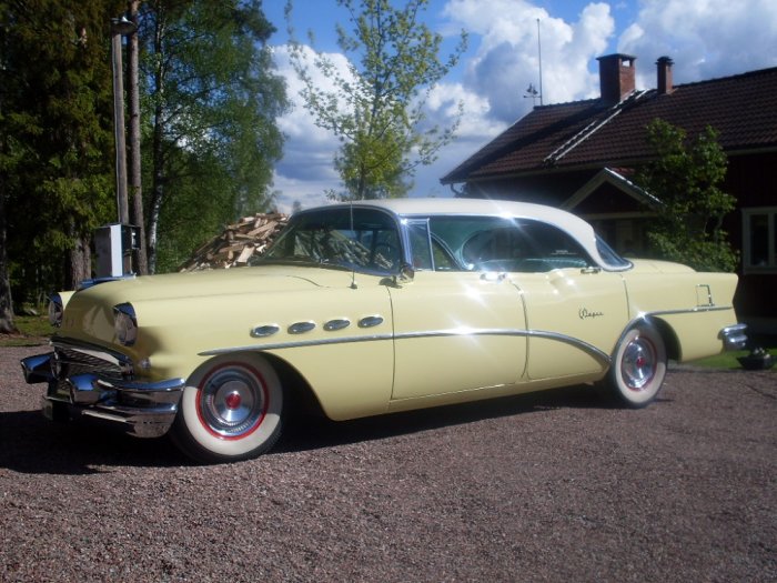 Gul och vit Buick Super Riviera från 1956 parkerad framför ett hus med vedstapel i bakgrunden.