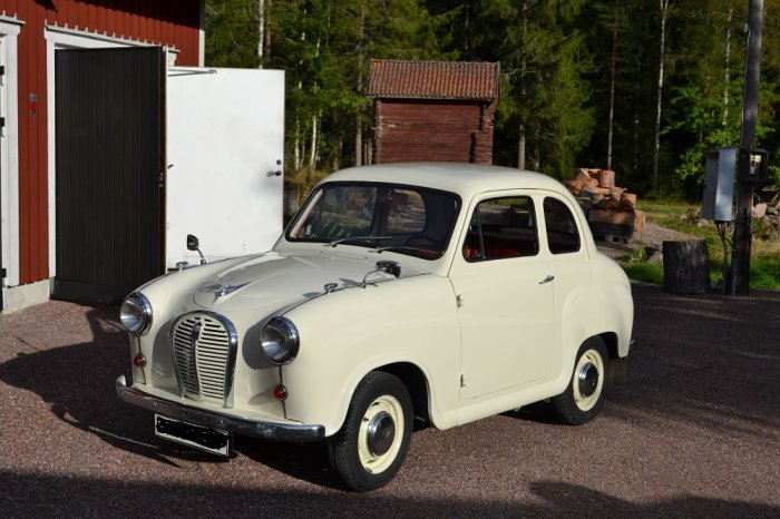 En krämvit Austin A35 från 1958 parkerad framför en röd lada och vita dörrar, omgiven av träd och vedstack.