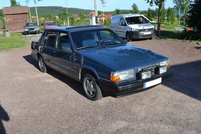 Blå Volvo 740 GLT från 1990 parkerad på grus med andra bilar i bakgrunden.