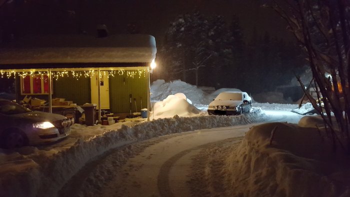 Vinterkväll med snötäckta bilar och väg, julbelysning på hus och snöskottad uppfart.