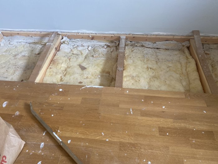 Golv med öppna spår där äldre gul isolation syns mellan träbjälkar.