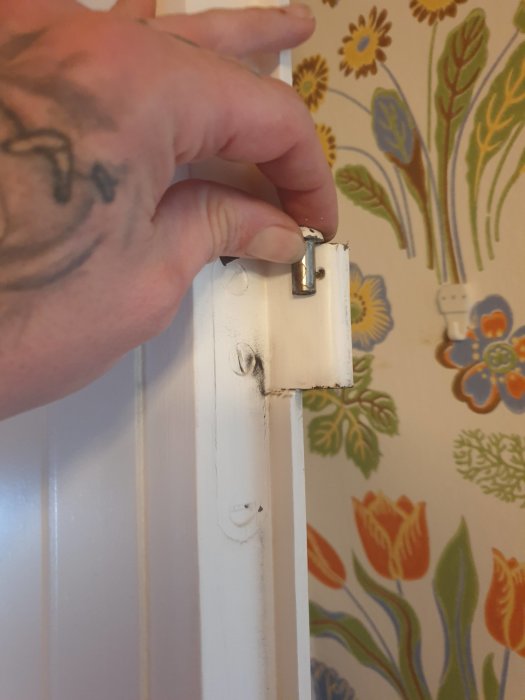 Hand som håller den avtagbara delen av en dörrs gångjärn framför tapet med blommönster.
