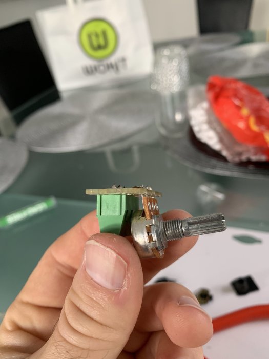 Hand håller en lodad elektronisk komponent med synliga lödningar och terminaler, otydlig bakgrund.